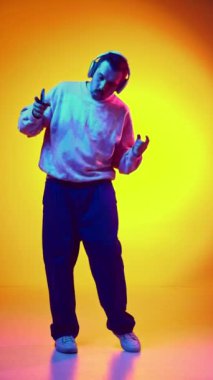 Pozitif, enerjik, pembe kazaklı ve kot pantolonlu, kulaklıkla müzik dinleyen ve neon rengi turuncu arka planda dans eden bir adam. İnsan duyguları kavramı, gündelik moda