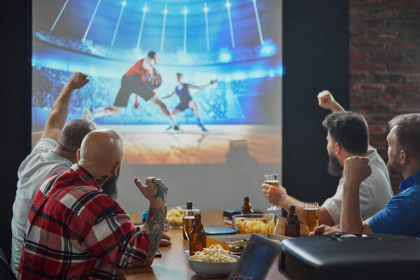 Men, friends sitting at table with beer and snacks, watching online basketball game translation on TV and cheering up favorite team. Concept of sport, championship, game, sport fans, leisure