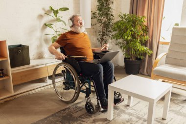 Oturma odasında, kulaklıkla tekerlekli sandalyede oturan ve dizüstü bilgisayardan davul çalmayı öğrenen yaşlı bir adam. Sağlık, yaşam tarzı, refah, rahatlık, güçlenme kavramı