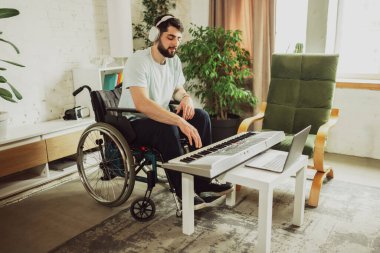 Oturma odasında tekerlekli sandalyede oturan ve dizüstü bilgisayardan piyano çalmayı öğrenen genç bir adam. Sağlık, yaşam tarzı, refah, rahatlık, güçlenme kavramı