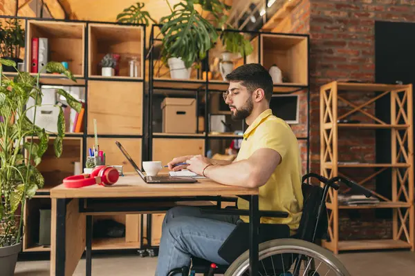 Young man, employee sitting on wheelchair and working remotely on laptop at home, Remote job, freelance. Concept of healthcare, lifestyle, wellness, comfort, empowerment