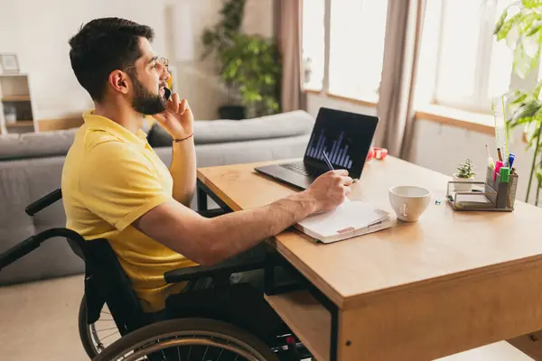 Businessman, employee, young man sitting on wheelchair at home, working online on laptop with financial analytics, talking on phone. Trading. Concept of healthcare, lifestyle, wellness, empowerment