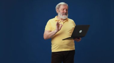 Son sınıf öğrencisi gülümsüyor ve mavi arka planda dizüstü bilgisayarla çevrimiçi video görüşmesi yapıyor. Uzaktan çalışma, çalışanlarla iletişim. İnsan duyguları kavramı, yaşam tarzı, gündelik moda.