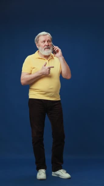 青いスタジオのバックグラウンドで携帯電話で話すカジュアルな服の笑顔 シニア ひげ付きの男の全長 通信する 人間の感情 ライフスタイル カジュアルなファッション テクノロジー — ストック動画