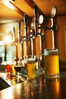 Ahşap tezgahta parlak metal bira muslukları. Muslukların yanında köpüklü bira kupası ve cam üzerinde yoğunlaşma var. İçecek bir şeyler. Bira, alkol, bira fabrikası, bar atmosferi, tat.