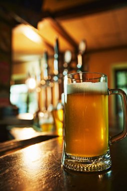 Arka plandaki rahat bar atmosferinde, ahşap masanın üzerinde köpüklü bira bardağı. Ferahlatıcı ve tatmin edici bir içecek. Bira, alkol, bira fabrikası, bar atmosferi, tat.