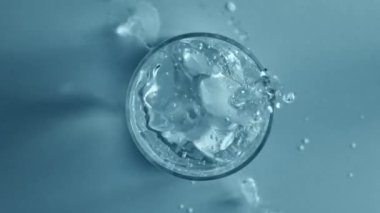Mavi zemin üzerinde izole edilmiş şeffaf, berrak su ile buzun cama düşüşünün üst görüntüsü. Susuzluğun temel rolü. Ferahlatıcı kavram, ürün estetiği, sağlık, sağlık