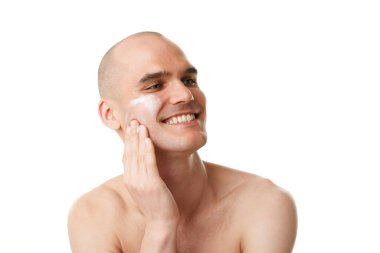 Beyaz stüdyo arka planına izole edilmiş nemlendirici krem süren lekesiz, bakımlı, yakışıklı genç bir adam. Cilt bakımı, erkek güzelliği, sağlık, kozmetik, kişisel bakım