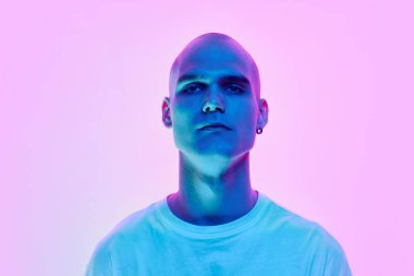 Ciddi bir genç adamın portresi, kel kafalı, yüzü ve kulağı traşlı, neon ışıklarıyla pembe arka planda poz veren. Erkek güzelliği, vücut, gençlik, spor, sağlık kavramı