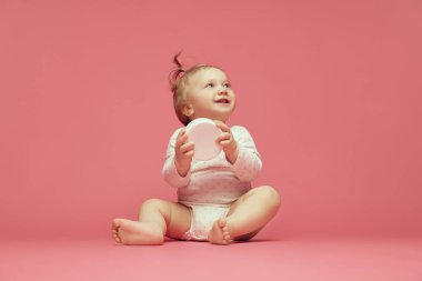 Pembe arka planda oturan rahat tulumlu gülümseyen bebek nemlendirici cilt kremi tutuyor. Bebekler için makyaj malzemeleri. Çocukluk kavramı, bakım, sağlık, refah, ebeveynlik