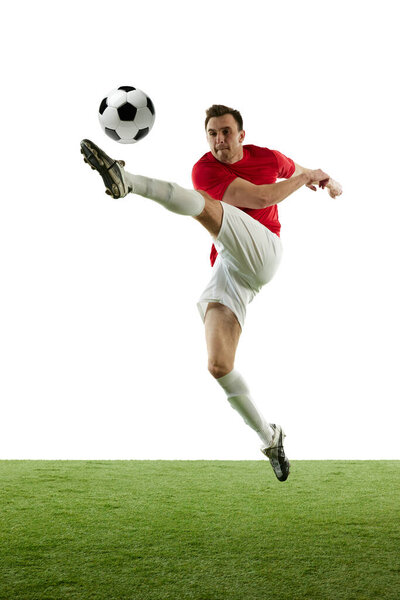 Full-length of young man in red and white uniform, football player hitting ball in a jump, training isolated on white background. Concept of professional sport, game, competition, tournament, action