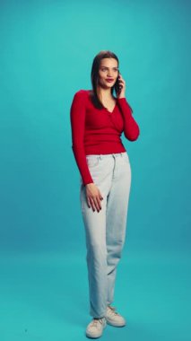 Kırmızı bluzlu ve mavi kotlu güzel bir kadın cep telefonuyla mavi stüdyo arka planıyla konuşuyor. İnsan duyguları kavramı, yüz ifadesi, yaşam tarzı, iletişim