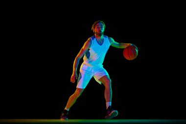 Genç atletik adam, top oynayan basketbol oyuncusu, neon ışıkta siyah arka planda antrenman yapıyor. Spor anlayışı, rekabet, aktif ve sağlıklı yaşam tarzı, oyun