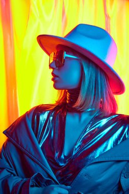 Gece partisi için hazırlan. Moda kıyafetleri, şapka, güneş gözlüğü ve parlak sarı arka planda poz veren şık bir genç kız. Modern moda kavramı, trendler, güzellik, gençlik kültürü