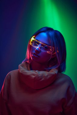 Modern rahatlık ve tarz. Yenilikçi gözlüklü rahat kapüşonlu genç kız neon ışıkta yeşil arka planda. Modern moda kavramı, moda tarzı, güzellik, gençlik kültürü