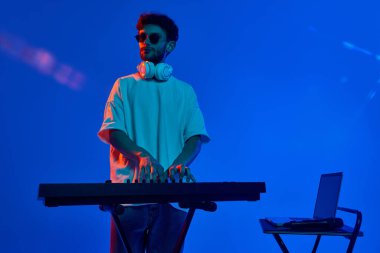Günlük kıyafetler, güneş gözlüğü ve kulaklıklar takan şık genç adam, neon ışıkta mavi arka planda DJ mikseriyle dinamik ritimler yapıyor. Müzik, performans, festival, konser, gece kulübü kavramı