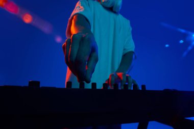 Genç erkek müzisyenin DJ mikseriyle birlikte çalması, farklı şarkıları neon ışıkta mavi arka planla karıştırması. Müzik, performans, festival, konser kavramı