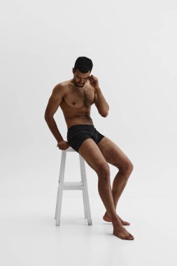 Beyaz arka planda izole edilmiş boxerlı, sandalyede üstsüz oturan yakışıklı Afro-Amerikan sakallı adamın tam boy görüntüsü. Erkek güzelliği, spor, vücut bakımı, sağlık, spor