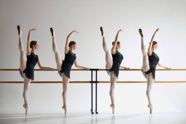 Genç kızlar, bale dansçıları zarafet ve esneklik gösteriyorlar, barre ve eğitimin yanında duruyorlar, gri stüdyo geçmişine karşı alıştırma yapıyorlar. Bale, sanat, dans stüdyosu, klasik tarz, gençlik kavramı.