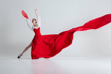 Flamenko sanatını icra eden canlı kırmızı elbiseli ve yelpazeli kadın dansçı gri stüdyo arka planına karşı zarafetle flamenko yapıyor. Hareket sanatı, klasik dans, güzellik kavramı