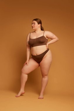 Güzel bir genç kızın tam boy görüntüsü, kendine güveni tam, artı beden, açık kahverengi arka planda iç çamaşırlarıyla duruyor. Doğal güzellik kavramı, vücut pozitifliği, özen.