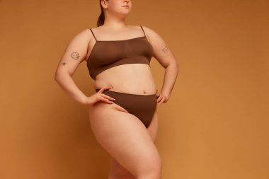 Pamuk iç çamaşırlarıyla açık kahverengi stüdyo arka planında poz veren iri vücutlu güzel bir genç kız görüntüsü. Doğal güzellik kavramı, vücut pozitifliği, özen, kabullenme