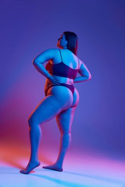 İç çamaşırlarıyla poz veren genç bir kızın tam boy arka plan görüntüsü. Mor renkli stüdyo ışıklarıyla büyük beden şekline sahip. Doğal güzellik kavramı, vücut pozitifliği, özen, kabullenme