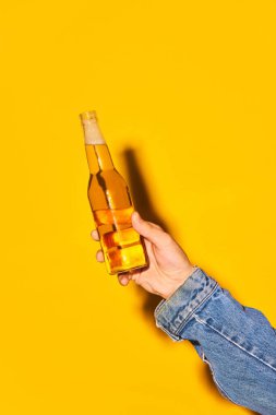 Parlak sarı arka planda bira şişesiyle insan eli tutmak. Alkol ve alkolsüz içecek kavramı, parti, tatil, bar, karışım. Poster. İlan için alanı kopyala