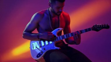 Yakışıklı, Afro-Amerikalı, müzikal oturan ve elektro gitar çalan neon ışıkta renkli stüdyo arka planına karşı. Müzik kavramı, canlı performans, sanat, yetenek, gösteri