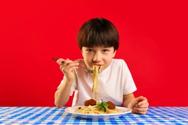 Lezzetli yemek zamanı. Küçük çocuk, çocuk, masa örtüsü ve kareli masa örtüsüyle oturuyor ve kırmızı arka planda köfteli spagetti yiyor. Yemek, çocukluk, duygular, yemek, pop sanat kavramı