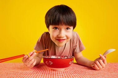 Yemek çubuğu ve kaşık kullanan neşeli genç bir çocuk, masada oturup ramen yiyor, sarı arka planda erişte çorbası içiyor. Yemek kavramı, çocukluk, duygular, Asya yemeği, menü, pop sanat