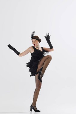 Tarz sahibi, genç, güzel bir kadın 1920 'lerin temalı kıyafetleriyle beyaz stüdyo arka planında neşeli bir şekilde dans ediyor. Sanat anlayışı, retro ve vintage, hobi, eğlence, 20 'ler