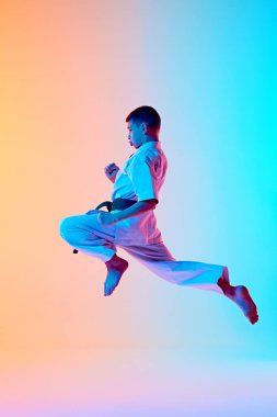 Young athlete engaged in karate training, jumping in dynamic pose against gradient orange blue background in neon light. Concept of sport, martial arts, combat sport, healthy and active lifestyle clipart