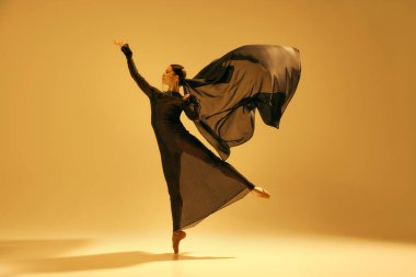 Güzel genç bir kadın, akışkan siyah elbiseli bir balerin altın sarısı arka planda gösteri yapıyor. Sanat kavramı, klasik dans, güzellik ve moda, estetik.