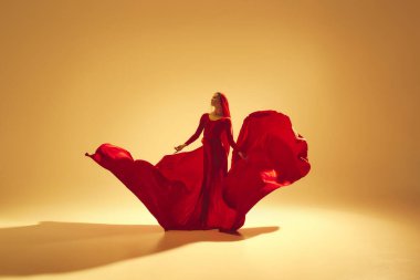 Zarif kırmızı akıcı elbiseli sanatçı kadın yaratıcı bir performans sergiliyor, altın renkli arka planda dans ediyor. Sanat kavramı, klasik dans, güzellik ve moda, estetik.