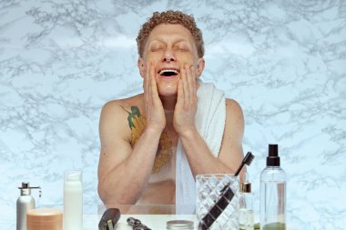 Omzunda havlu olan gömleksiz bir adam sabah banyoda rutinini yapıyor, yüzünü yıkıyor. Ön planda çeşitli deri ve diş ürünleri var. Erkek güzelliği, cilt bakımı, kozmetik ve kozmetik kavramı