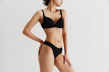 Kusursuz ince kadın vücudunun kırpılmış görüntüsü. Gri stüdyo arka planında siyah iç çamaşırlarıyla poz veren bir model. Vücut pozitifliği. Doğal güzellik kavramı, cilt bakımı, spor, sağlık, sağlık