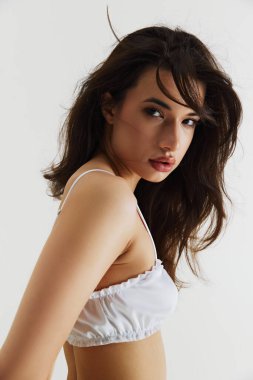 Gri stüdyo arka planında izole edilmiş beyaz zarif iç çamaşırlarıyla poz veren tombul dudaklı güzel esmer kız portresi. Doğal güzellik kavramı, cilt bakımı, spor, sağlık, sağlık