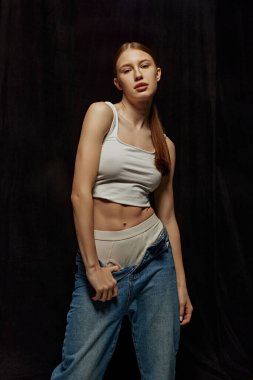 İnce vücutlu, çıplak makyajlı, beyaz bluz ve kot pantolon giyen, siyah arka planda poz veren güzel bir genç kız. Güzellik, vücut, spor ve sağlık, moda, sağlık kavramı