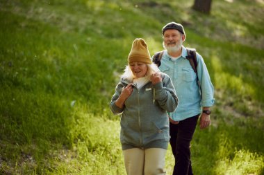 Mutlu, gülümseyen yaşlı bir çift, erkek ve kadın el ele tutuşuyor, güneşli bir günde parkta yürüyorlar. Yürüyüş aktivitesi. Spor anlayışı, yaşlanma, aktif ve sağlıklı yaşam tarzı, sağlık hizmetleri