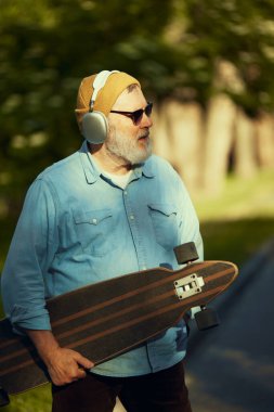 Gündelik giysiler içinde, güneş gözlüğü, kulaklık takan ve kaykayla ayakta duran şık bir son sınıf öğrencisi. Aktif hobi. Spor anlayışı, yaşlanma, aktif ve sağlıklı yaşam tarzı, sağlık hizmetleri