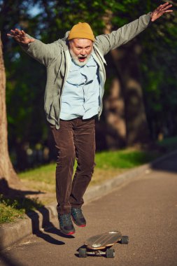 Güneşli bir günde, sabah parkında kaykay yapan, günlük şık kıyafetler giyen yaşlı bir adam. Gençlik aktiviteleri. Spor anlayışı, yaşlanma, aktif ve sağlıklı yaşam tarzı, sağlık hizmetleri