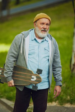 Düşünceli yaşlı adam kaykayını tutuyor, parkta dinlenmek için bir an duruyor. Aktif hobi ve iyi sürüş. Spor anlayışı, yaşlanma, aktif ve sağlıklı yaşam tarzı, sağlık hizmetleri