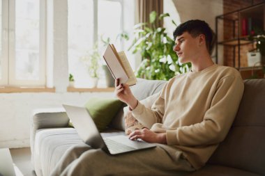 Gündelik giysiler içindeki genç adam evdeki modern odada oturuyor, kitap okuyor ve not almak için dizüstü bilgisayar kullanıyor. Edebiyat öğrencisi. Eğitim, öğrenim, ödev, gençlik, yaşam tarzı kavramı