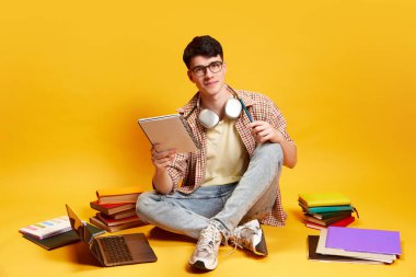 Genç, gülümseyen bir adam, gündelik giysiler içinde erkek öğrenci bir sürü kitabın, defterlerin ve dizüstü bilgisayarın etrafında parlak sarı arka planda oturuyor. Eğitim, öğrenim, ödev, gençlik, yaşam tarzı kavramı