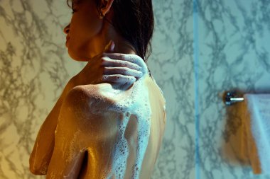 Kadın rahatlatıcı bir duş alıyor, vücudu kokulu duş jeliyle yıkıyor, vücutta köpük var. Neon ışıklı mermer banyo. Güzellik, kaplıca, rahatlama, banyo kozmetik ürünleri, sağlık.