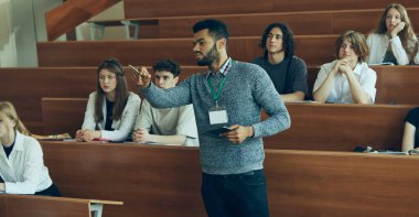 Genç öğretmen, üniversite konferans salonunda ders veren öğretmen, öğrencilere ilgi gösteren, tahtaya işaret eden. Sınıfta çok ırklı öğrenciler var. Eğitim kavramı, gençlik, atölye, bilgi