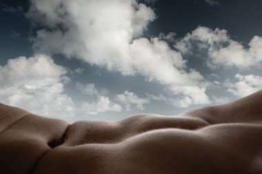 Bulut oluşumlarının güzelliği ve insan vücudu kıvrımları. Bronzlaşmış erkek kaslı vücudunun yakın çekimi arka planda bulutlu gökyüzü ile doğal bir manzara oluşturuyor. Vücut estetiği, insan doğası ve güzelliği kavramı