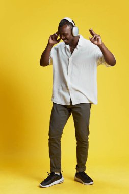 Gündelik giysiler içinde Afro-Amerikan bir adamın tam boy portresi kulaklıkla müzik dinliyor ve parlak sarı arka planda dans ediyor. İnsan duyguları kavramı yüz ifadesi, moda