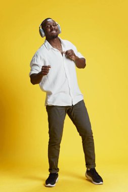 Tam boy Afro-Amerikalı şık günlük kıyafetler içinde kulaklıkla müzik dinleyen ve parlak sarı arka planda gülümseyerek dans eden bir adam. İnsan duyguları yüz ifadesi, moda konsepti
